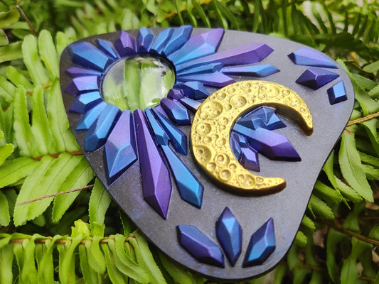 Decorative planchette with blue and purple crystal design and gold crescent moon on green leaves