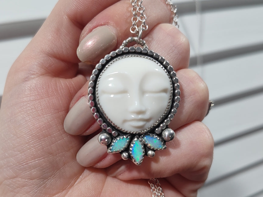 A handcrafted silver pendant with white porcelain moon face and 3 marquise shaped opals on a silver chain, held in hand for scale.