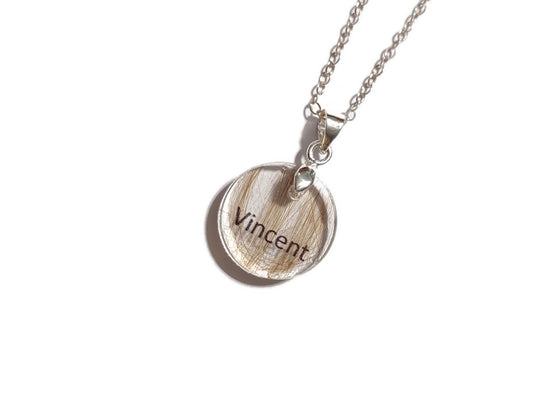 Clear necklace with a round pendant with 'Vincent' and a small amount of hair inside, on a white background