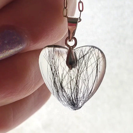 A small heart shaped keepsake pendant containing black hair, being held in a hand in front of natural light.