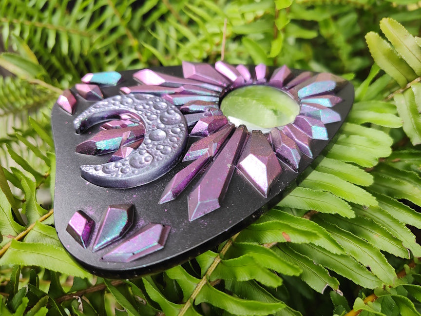 Decorative planchette with pink to blue chameleon crystal pattern and crescent moon on a green leaf 