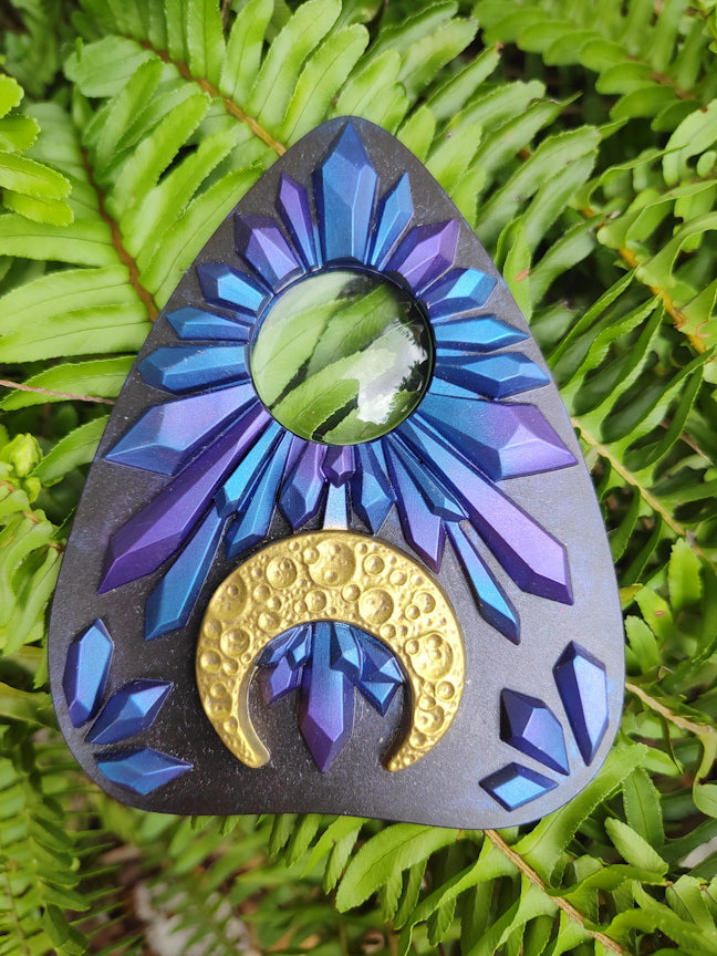Decorative planchette with blue and purple crystal design and gold crescent moon on green leaves