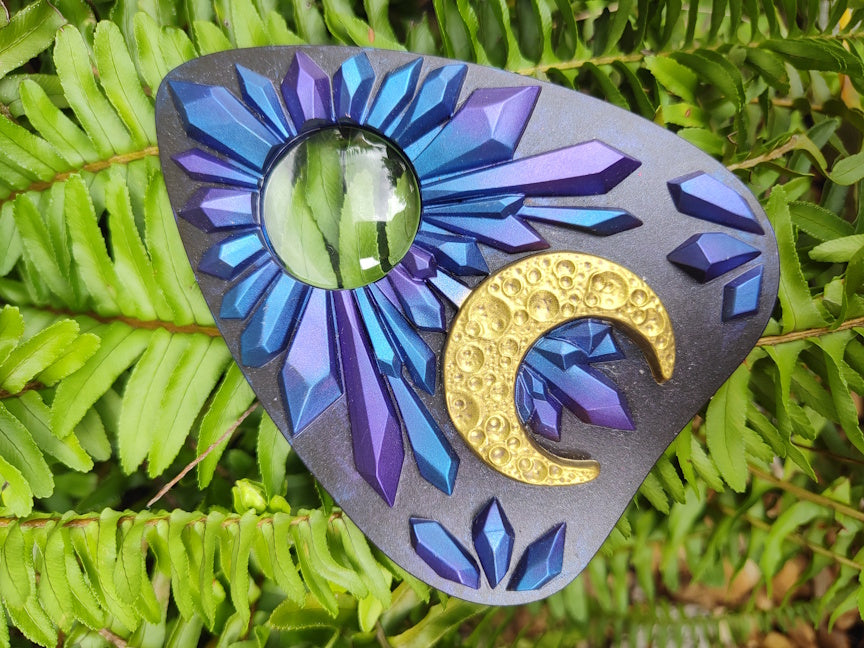 Decorative planchette with blue and purple crystal design and gold crescent moon on green leaves