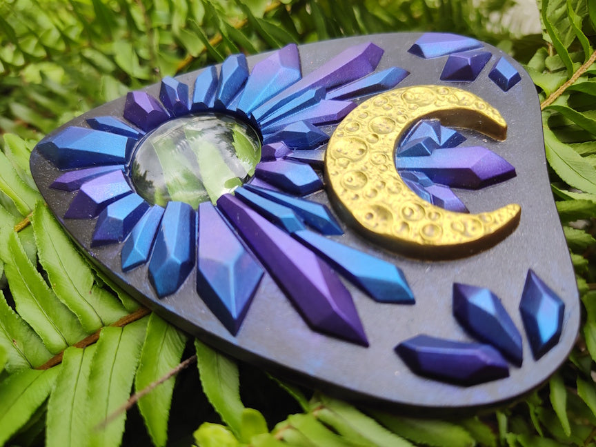 Decorative planchette with blue and purple crystal design and gold crescent moon on green leaves