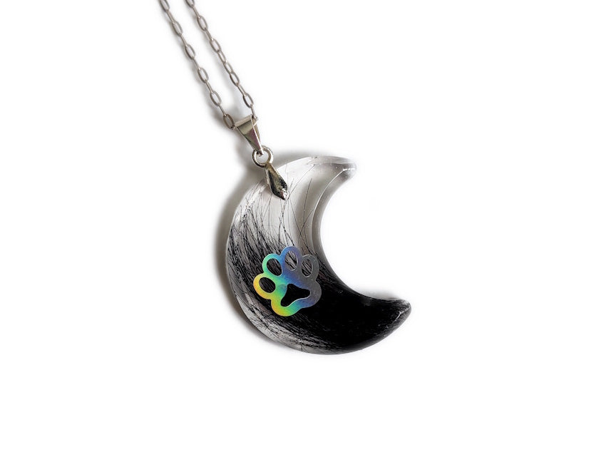 A moon shaped pendant with pet fur and holographic paw print, on a silver chain over a white background.