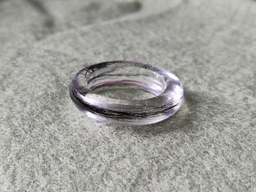A clear resin ring with black hair inlay, resting on a textured gray-green surface.