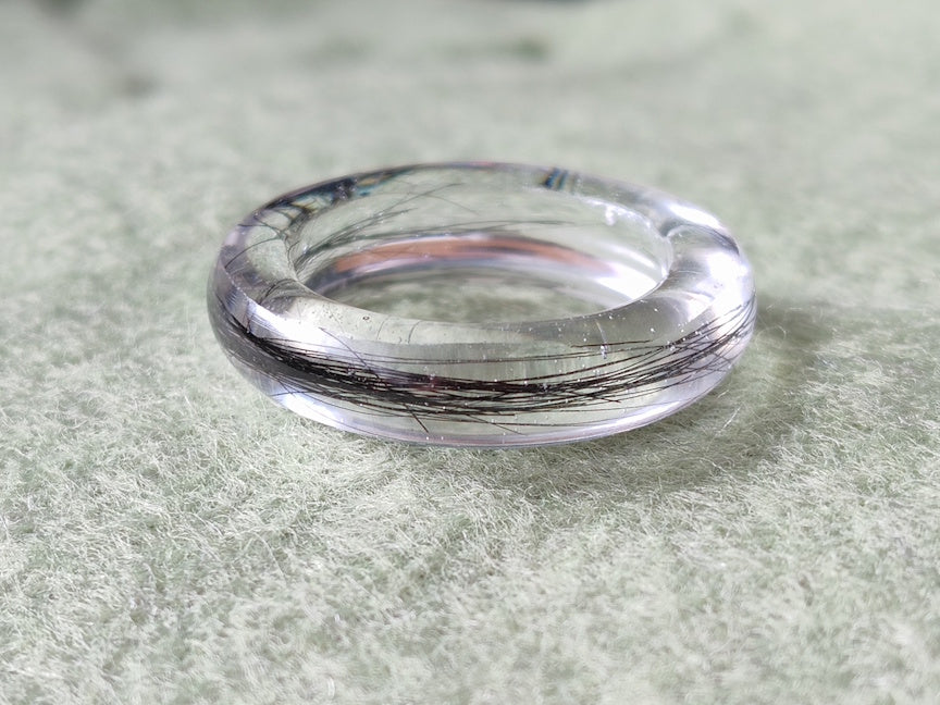 A clear resin ring band with visible dark hair inclusions, photographed on a soft light gray textured surface.