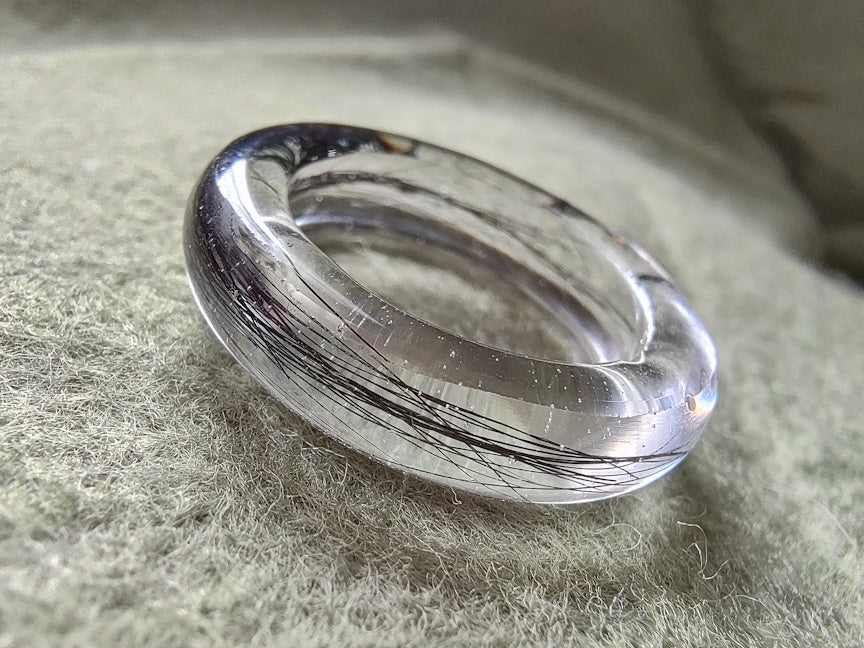 A clear resin ring with black hair inclusions, resting on a textured gray-green fabric background.