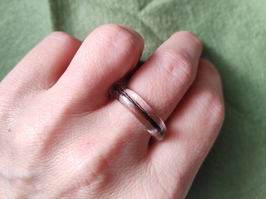 A close-up of a hand wearing a polished clear resin ring with black hair inside, set against a textured sage green background.