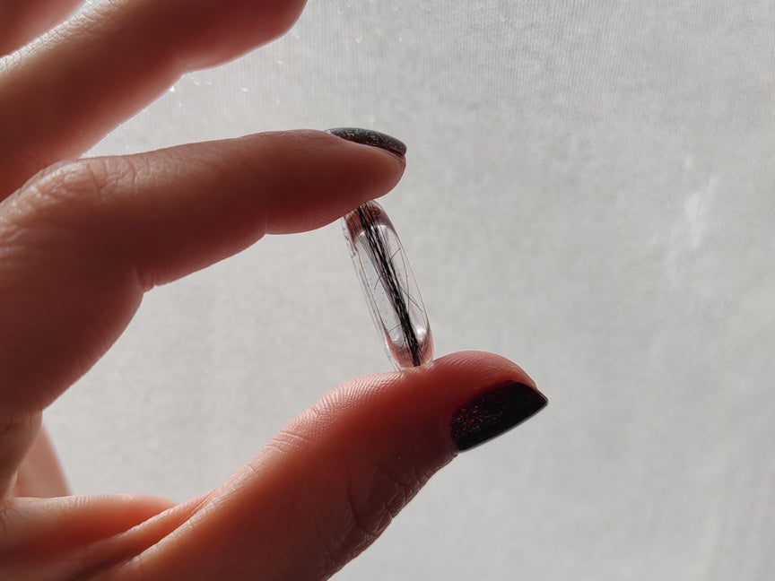 A close-up of a hand holding a clear resin ring containing hair, set against a soft, light gray textured background.
