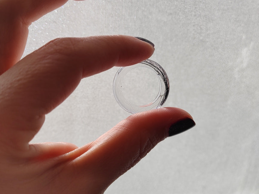 A close-up of a hand holding a clear, transparent ring against a soft, light gray textured background.