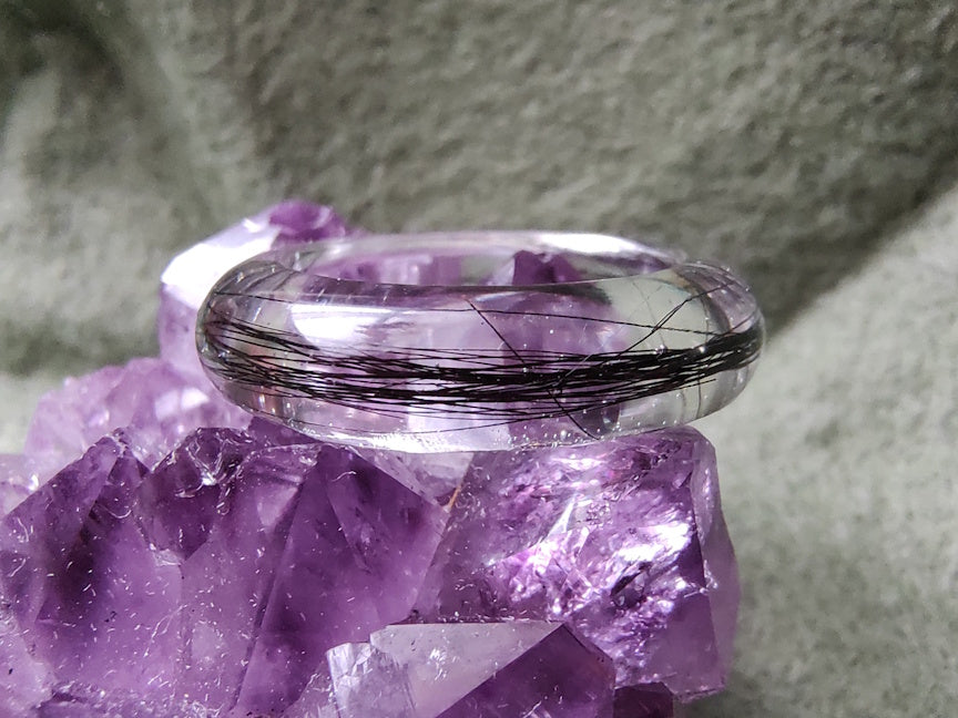 A close-up view of a polished clear resin ring with black hair preserved inside, on top of a purple crystal with green background.