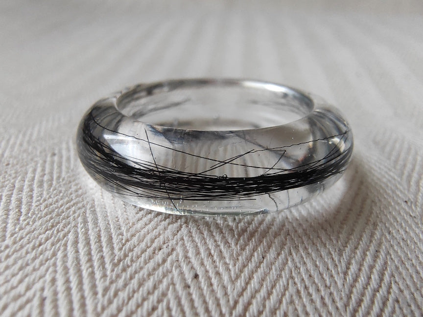 A clear resin ring with black hair inlay, photographed on a textured off-white fabric background.