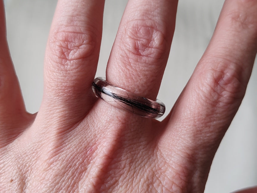 A close-up of a hand wearing a clear resin ring with black hair inlay, shown against a neutral light gray background.