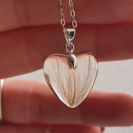 A small heart shaped keepsake pendant containing a small amount of light brown hair, being held in a hand in front of natural light.