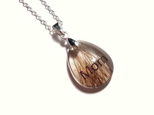 A small teardrop shaped pendant with hair and the name Mom. 
